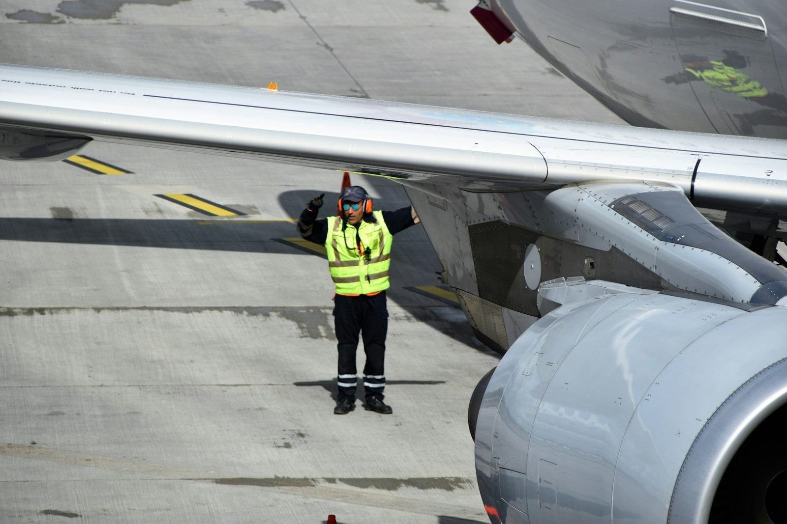 Eine Person in Warnweste und mit Gehörschutz steht unter der Tragfläche eines Flugzeugs und gibt ein Handzeichen
